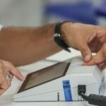 Mãos de um mesário e de um eleitor na identificação biométrica diante da urna eletrônica