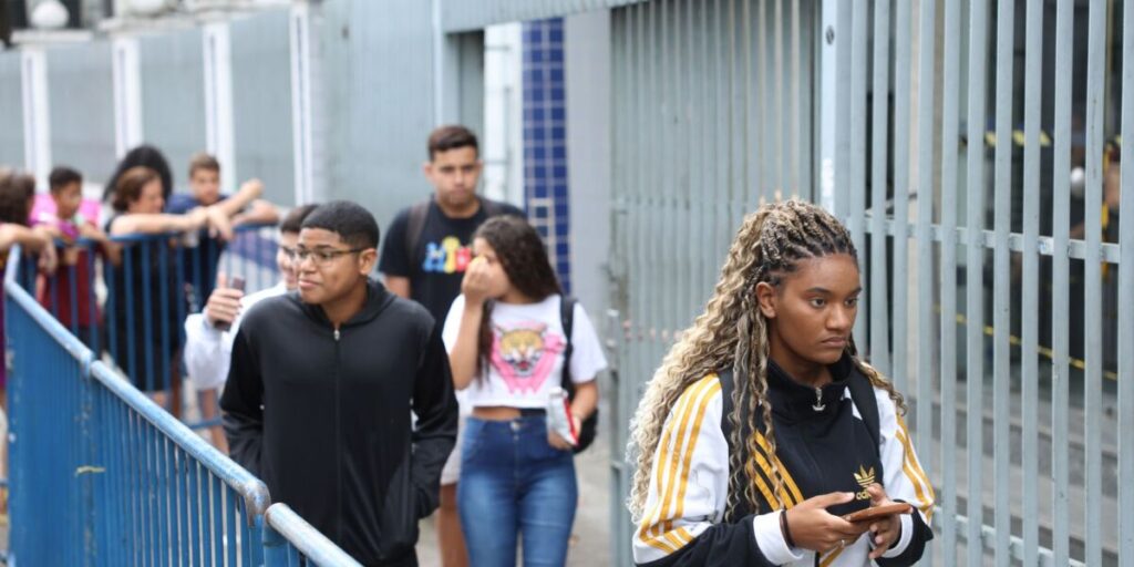 Estudantes caminham por uma calçada ao lado de um gradil, em frente a uma escola, em dia de prova
