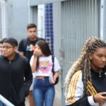 Estudantes caminham por uma calçada ao lado de um gradil, em frente a uma escola, em dia de prova