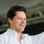 O governador de Goiás, Daniel Vilela, sorrindo e acenando com a mão levantada durante um evento, vestindo camisa branca