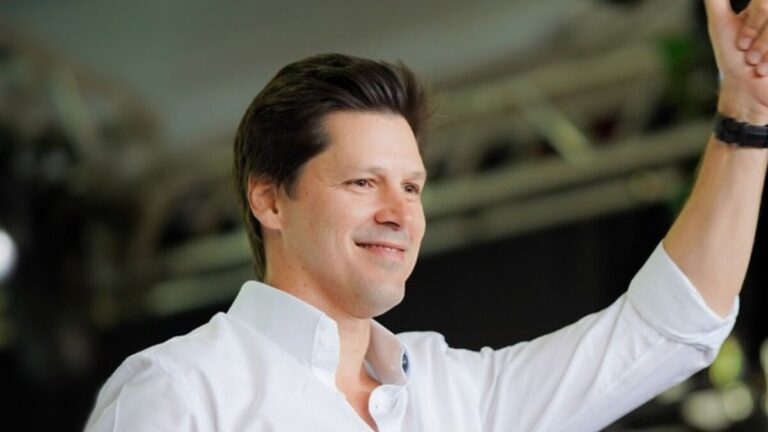 O governador de Goiás, Daniel Vilela, sorrindo e acenando com a mão levantada durante um evento, vestindo camisa branca