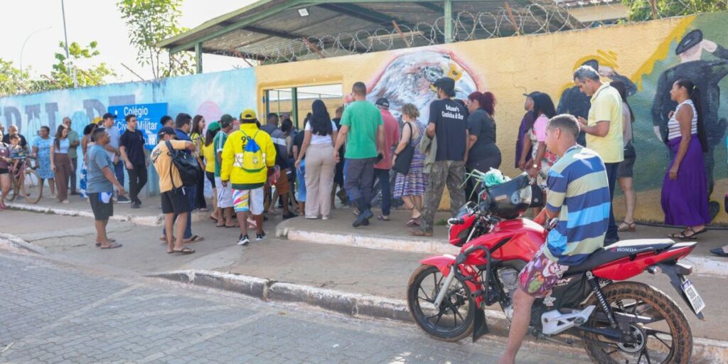 Eleitores em fila na calçada diante de uma escola com muro grafitado, em dia de votação, com um motociclista parado na frente