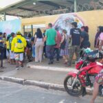 Eleitores em fila na calçada diante de uma escola com muro grafitado, em dia de votação, com um motociclista parado na frente