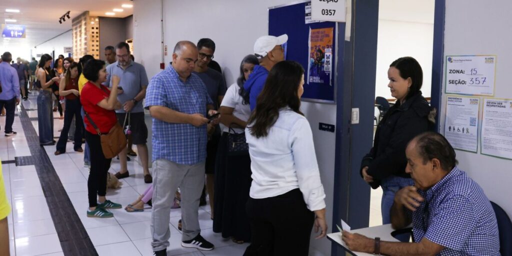 Fila de eleitores em um corredor, diante de salas de votação identificadas por número de zona e seção, em um colégio