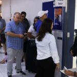 Fila de eleitores em um corredor, diante de salas de votação identificadas por número de zona e seção, em um colégio