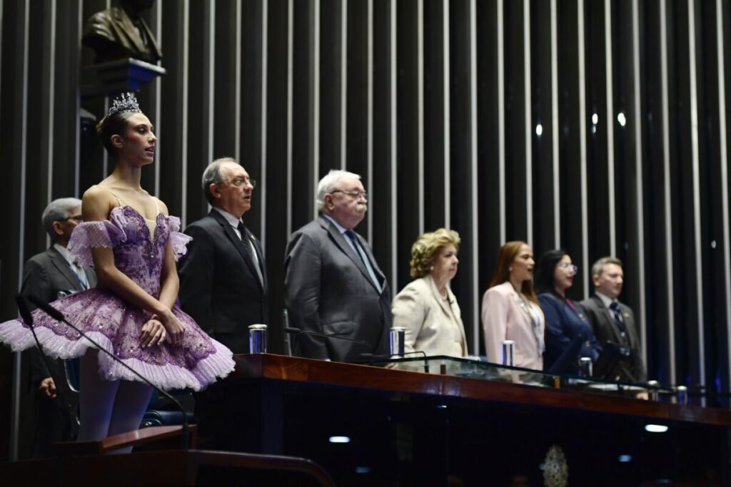 Bailarina de tutu roxo e coroa em pé, ao lado de autoridades em uma mesa, durante sessão especial no plenário do Senado