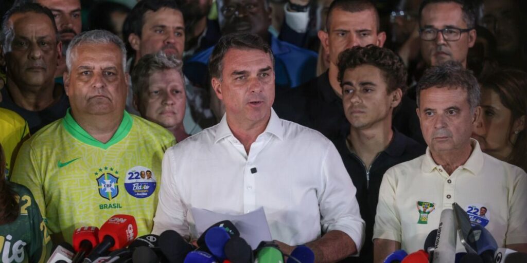 O senador Flávio Bolsonaro, de camisa branca, fala diante de vários microfones, cercado de aliados, durante pronunciamento