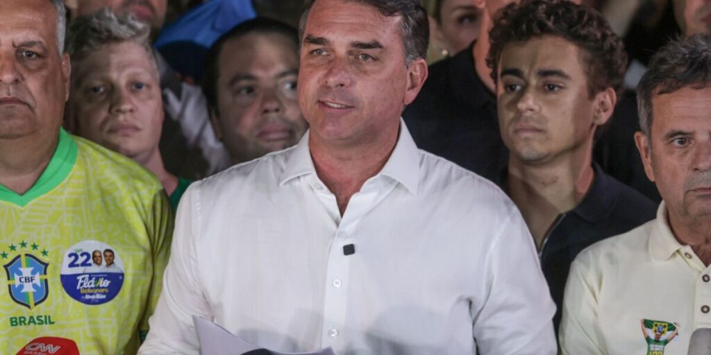 O senador Flávio Bolsonaro, de camisa branca, fala diante de aliados em pronunciamento, com camisetas amarelas ao redor