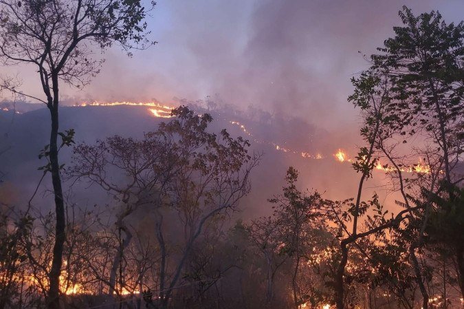 Linha de fogo avançando em uma encosta de vegetação de cerrado, com fumaça ao fundo