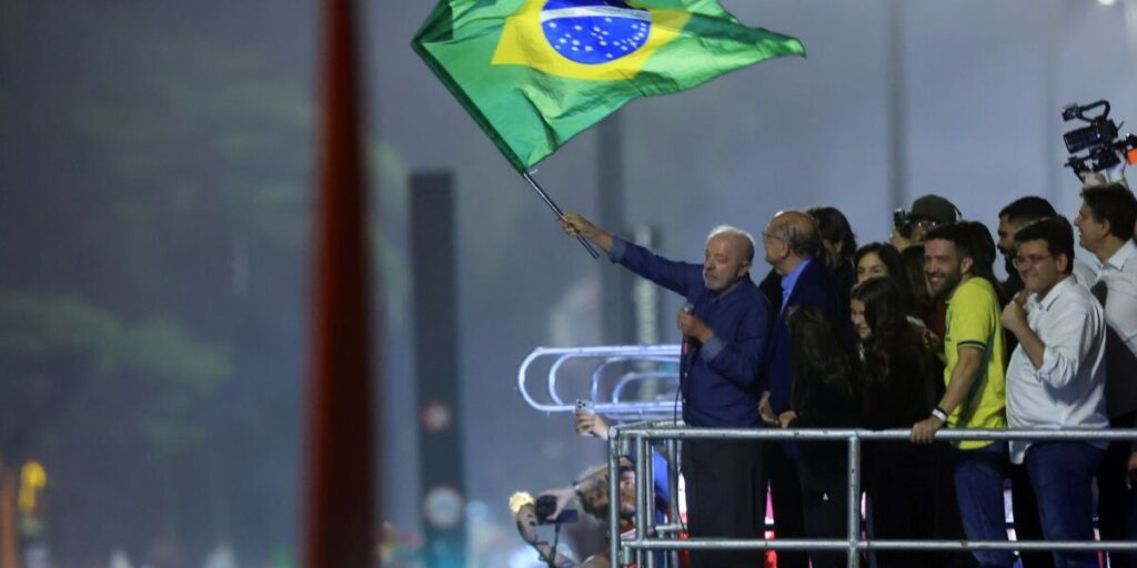 Lula acena com uma bandeira do Brasil em cima de um palanque, à noite, cercado de aliados, na Avenida Paulista