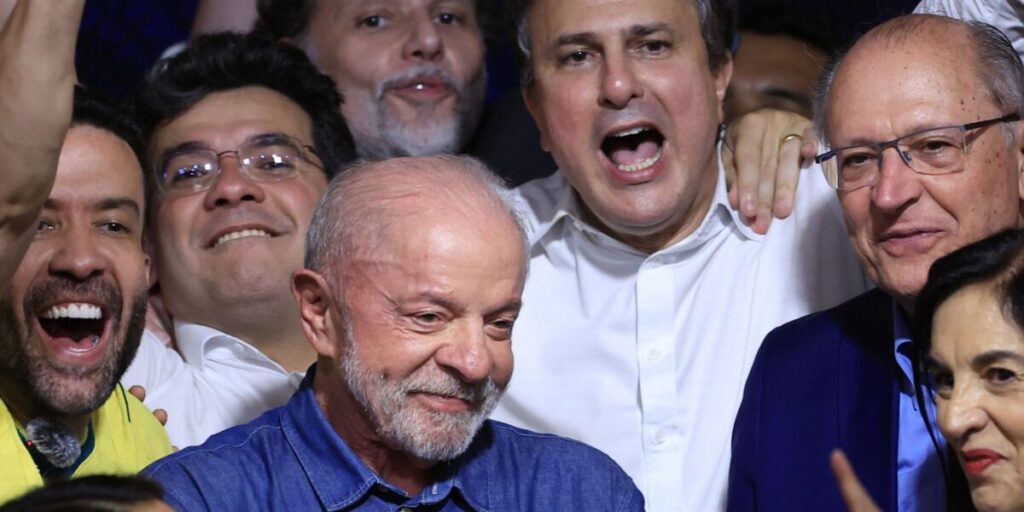 Lula sorri ao centro de um grupo de aliados que comemoram e cantam, em meio à festa da vitória