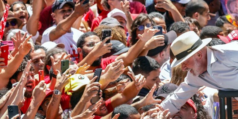 Lula, de chapéu claro, se inclina para cumprimentar apoiadores que erguem celulares e as mãos na festa da vitória