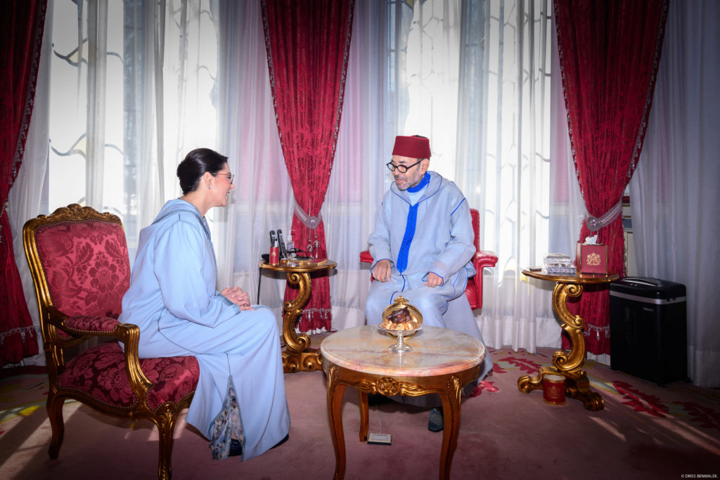 Rei Mohammed VI do Marrocos, de caftan azul-claro e gorro vermelho, sentado em frente a Fatima Ezzahra El Mansouri, também de caftan azul-claro, em salão de palácio com cortinas vermelhas e móveis dourados