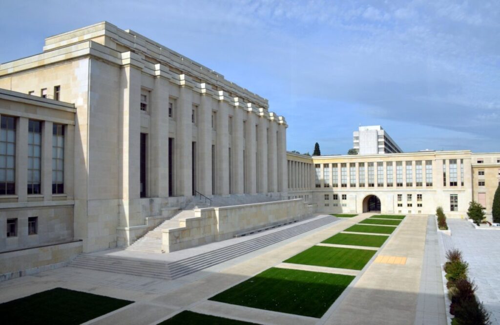 Fachada do Palácio das Nações, sede do escritório da ONU em Genebra, com bandeiras e jardim