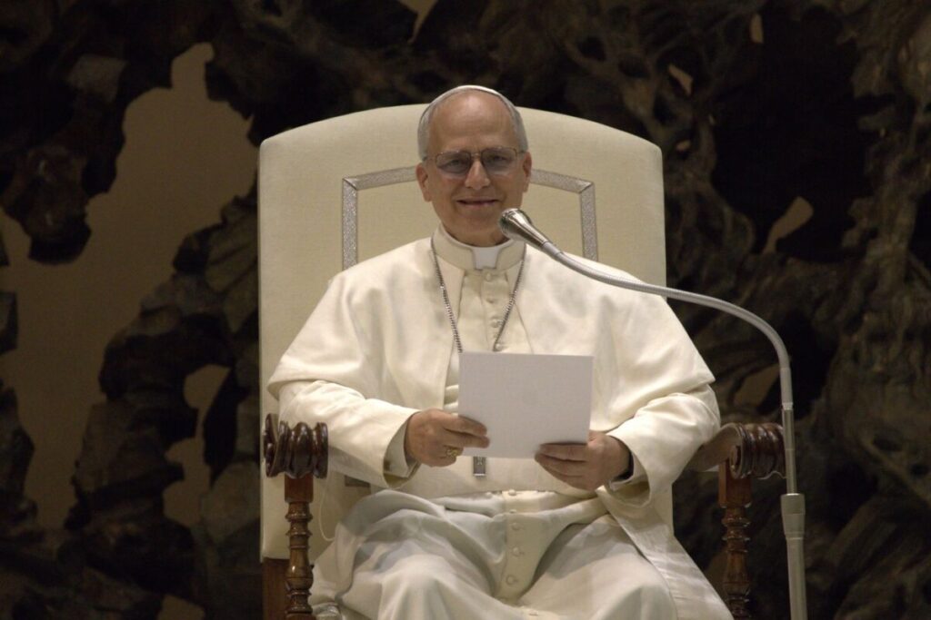 O papa Leão XIV sentado em uma cadeira branca, sorrindo e segurando uma folha, diante de um microfone