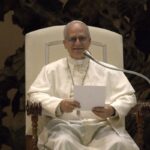 O papa Leão XIV sentado em uma cadeira branca, sorrindo e segurando uma folha, diante de um microfone
