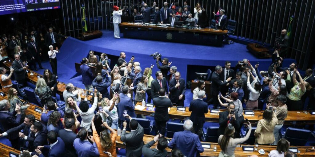 Plenário do Senado em momento de comemoração após a votação, com parlamentares de pé e fotógrafos no centro