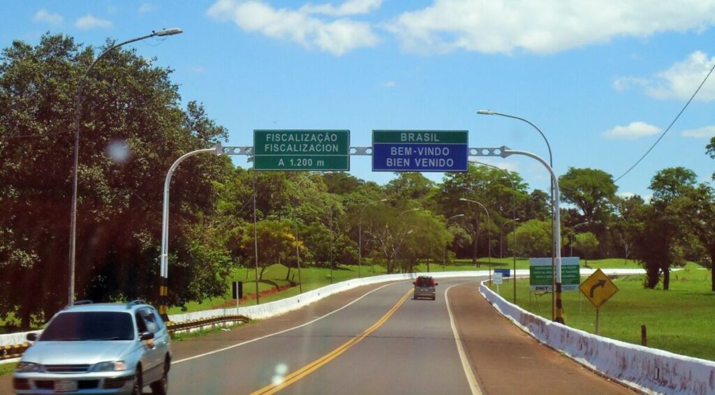 Pista de acesso a uma ponte internacional, com placas sobre a pista indicando Brasil, bem-vindo e fiscalização a 1.200 metros